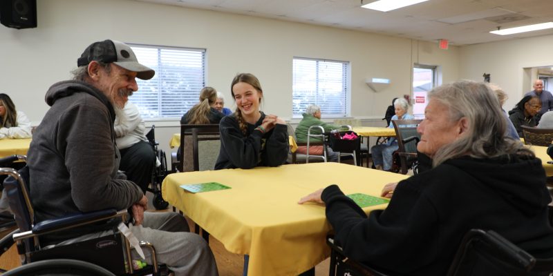 People sitting at tables in a brightly lit room, engaging in conversation. Some are in wheelchairs. Yellow tablecloths are on the tables.