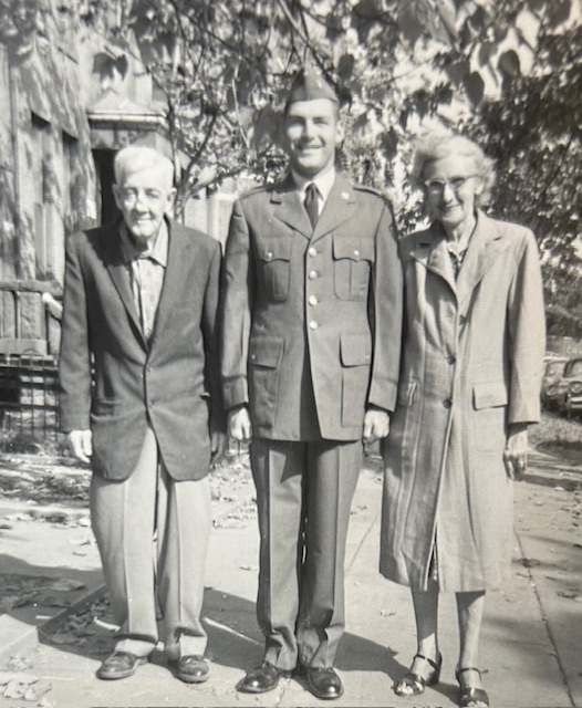Three people stand on a sidewalk under trees. A man in a military uniform is flanked by an elderly man on the left and an elderly woman on the right, all smiling at the camera.