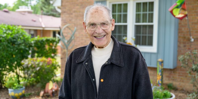 Elderly man smiling in a garden with a brick house in the background.