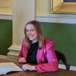 A person in a pink jacket sits at a wooden table strewn with papers and glasses, deep into their Political Science studies. Against a green and white wall with a large frame, they smile warmly at the camera, light brown hair softly falling over their dark top.
