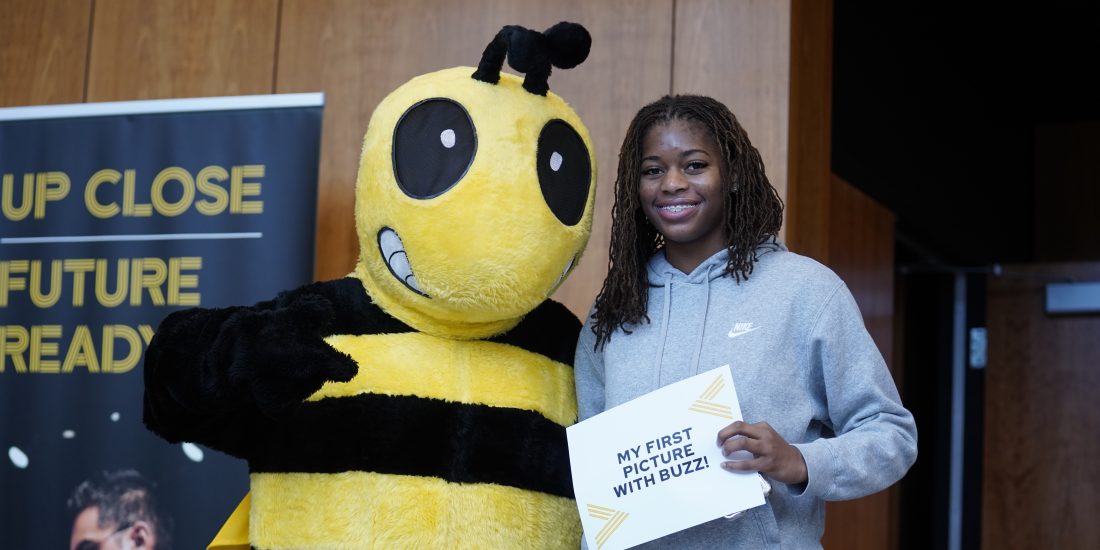 A student poses with someone in a Yellow Jacket costume, holding a sign that reads, "My first picture with Buzz."