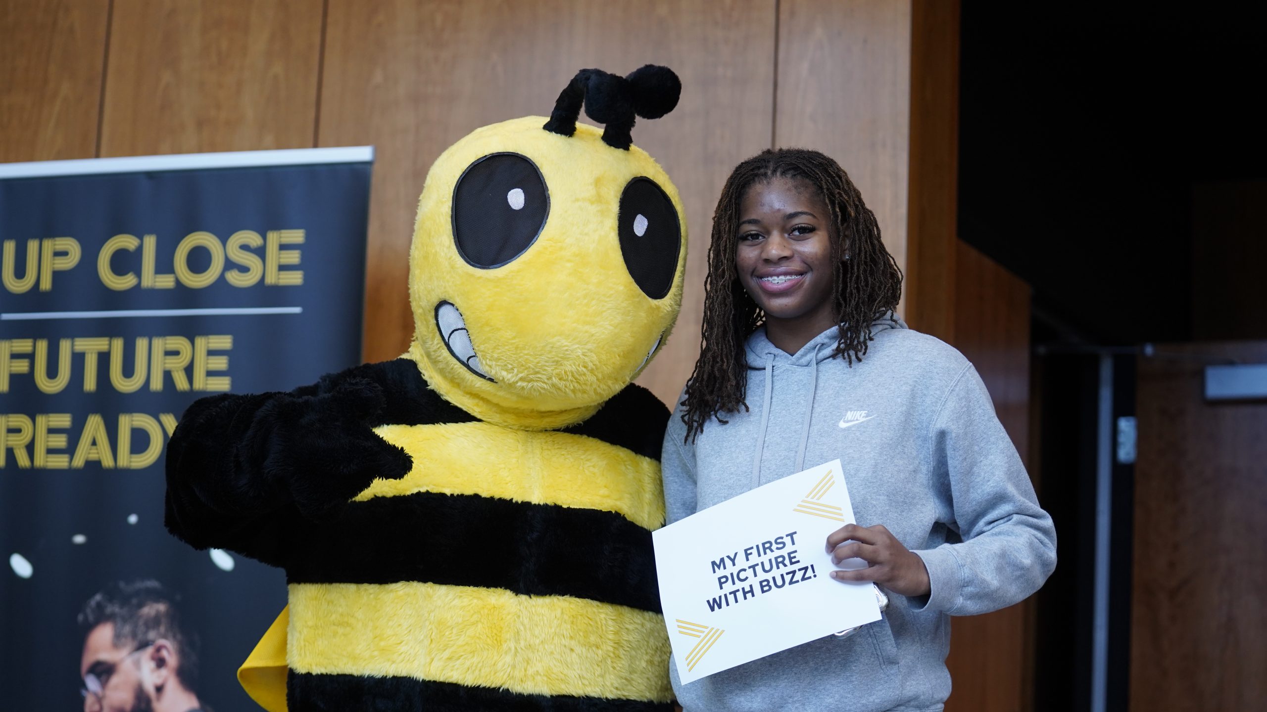 A student poses with someone in a Yellow Jacket costume, holding a sign that reads, "My first picture with Buzz."