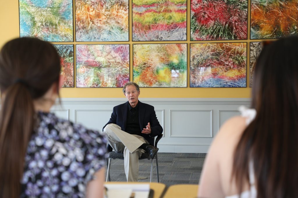 A person sits and talks to two others in a room with colorful abstract paintings on the wall.