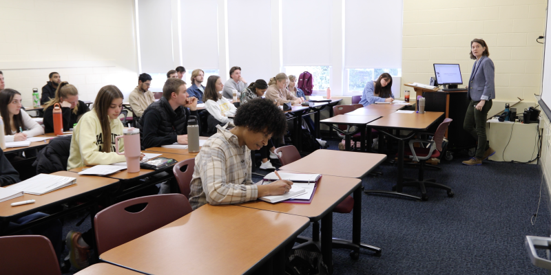 A teacher stands at the front of a classroom, presenting to students seated at desks, taking notes and listening attentively.