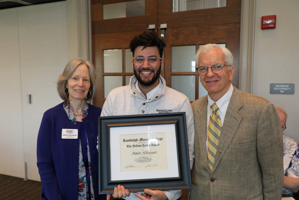 Three people stand indoors; a middle-aged woman, a smiling man holding a framed certificate, and an elderly man. The certificate reads "The Phi Beta Kappa" from Randolph-Macon College.