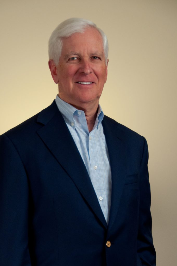 An older man with short white hair is wearing a dark blue blazer and light blue dress shirt, standing and smiling against a plain beige background.