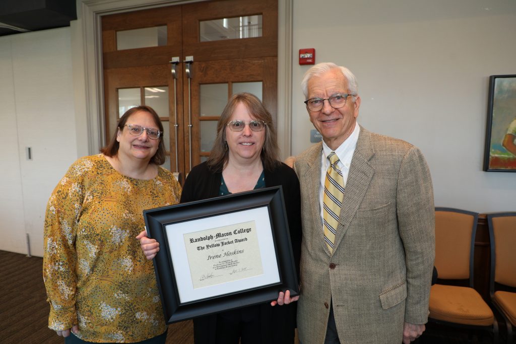 Three people pose indoors; the woman in the center holds a framed certificate.