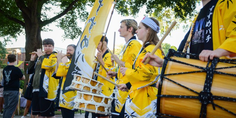 People in yellow jackets play traditional Japanese instruments outdoors, including flutes and drums, under a tree.
