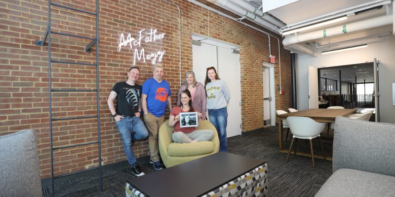 Five people pose together in a modern lounge with brick walls, a neon "McFather Mezz" sign, and contemporary furniture; one person is holding a black and white photo of a woman.