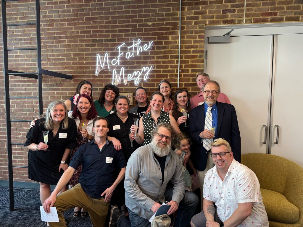 A group of people pose and smile together indoors in front of a brick wall with a neon sign that reads "McFather Mezz." Most are holding drinks and wearing name tags.