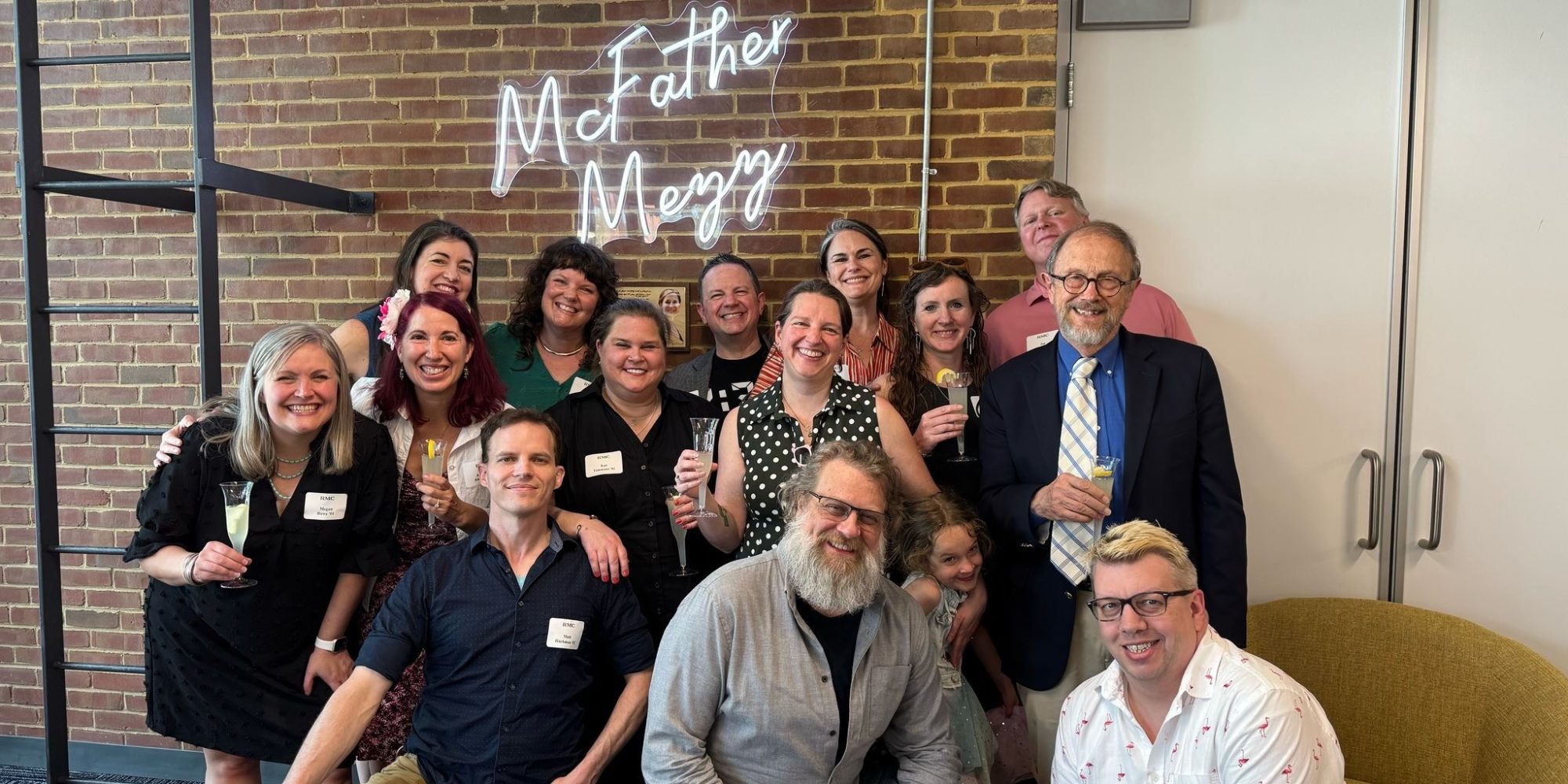 A group of people pose and smile together indoors in front of a brick wall with a neon sign that reads 