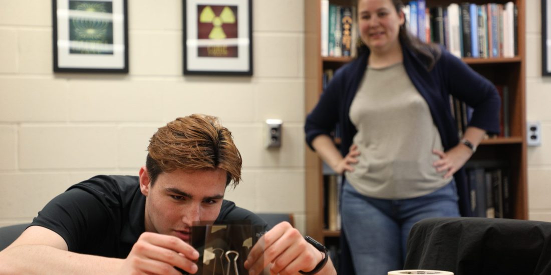 A person closely examines a polarizing filter on a table while another stands nearby in a room with framed artwork and a bookshelf.