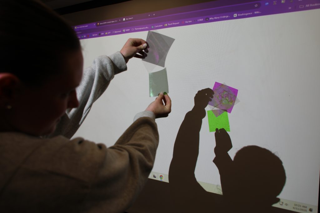 A person holds translucent colored sheets in front of a screen, casting shadows on it.