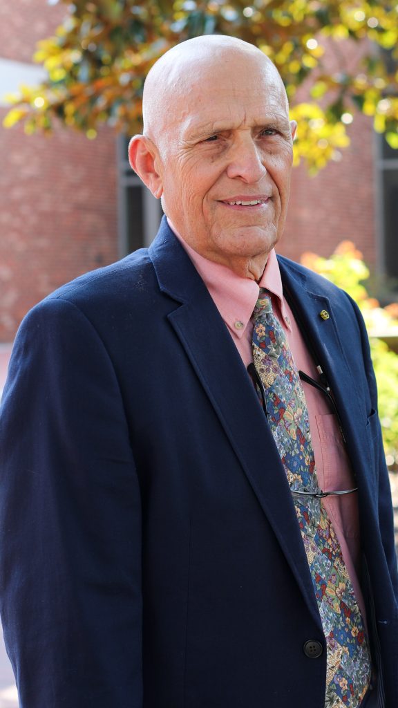 An older man wearing a navy suit, pink shirt, and patterned tie stands outdoors in front of a brick building and greenery.