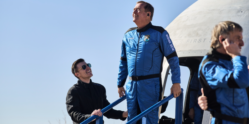 A person in a blue space suit is smiling at another person on the ground while standing on a set of stairs beside a spacecraft.