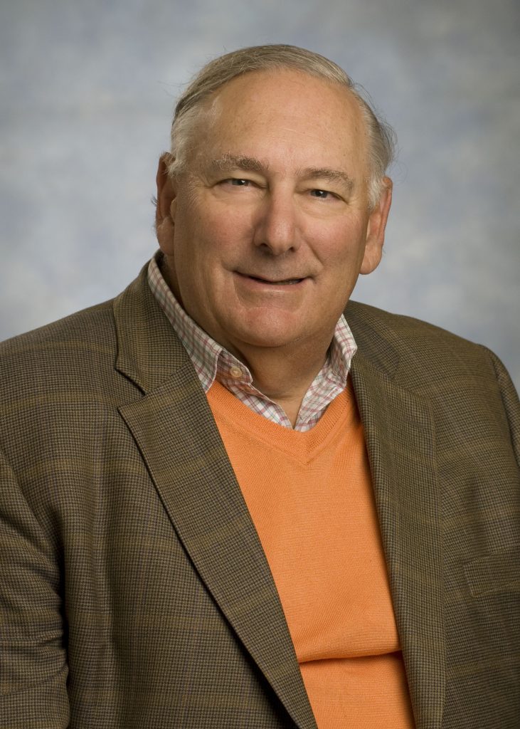 A middle-aged man with gray hair wearing a brown blazer, plaid shirt, and orange sweater poses for a portrait against a light blue background.
