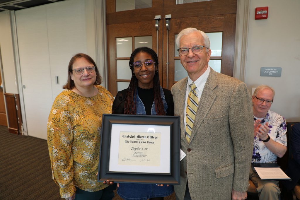 Three people pose with a framed certificate in a room. A woman on the right is clapping in the background.