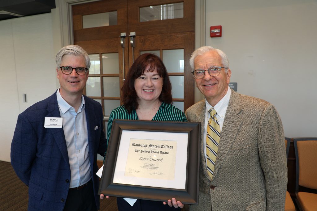 Three people smiling, with one in the center holding a framed award certificate, standing indoors in front of wooden doors.