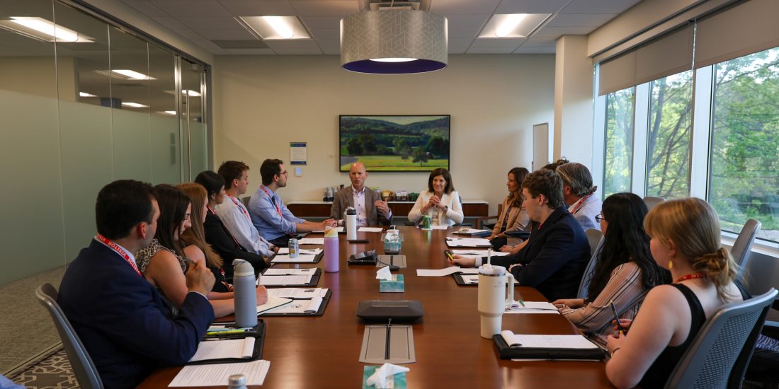 A group of people in business attire sit around a long conference table in a meeting room, listening to a person speaking at the head of the table.