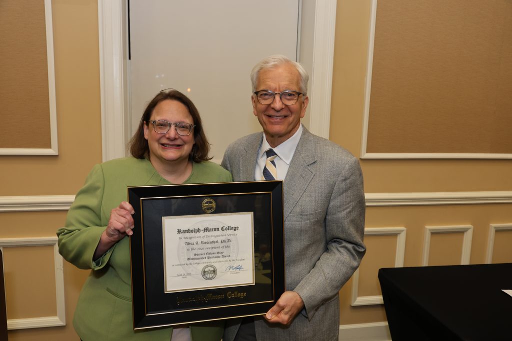 Two people stand indoors and smile at the camera while holding a framed Randolph-Macon College diploma or certificate together.