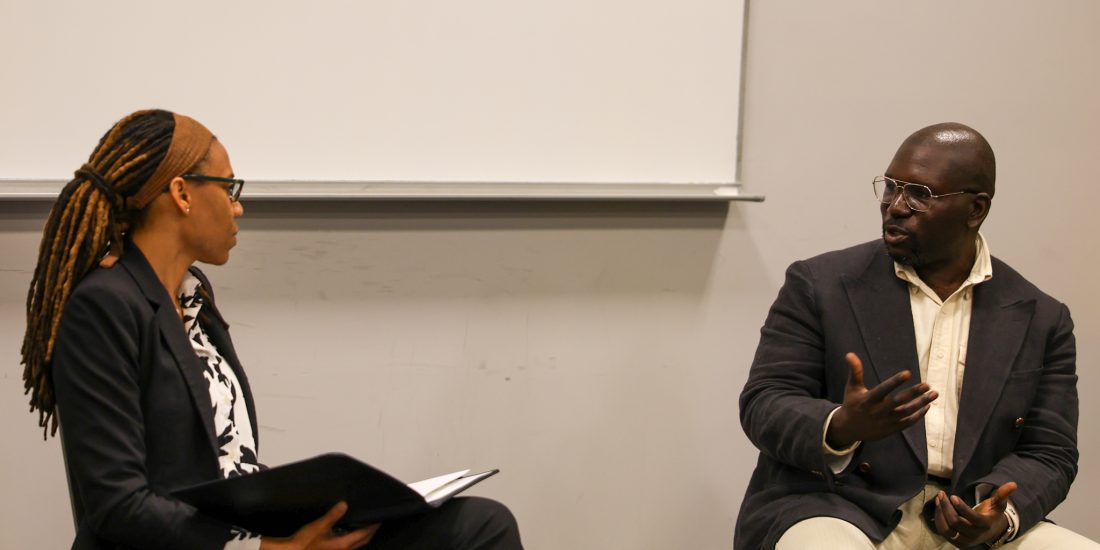 Two people in business attire are engaged in conversation in a classroom setting with a poster on the wall behind them.