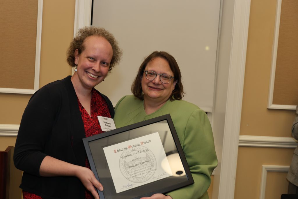 Two women stand indoors, one holding a framed certificate while both smile at the camera.