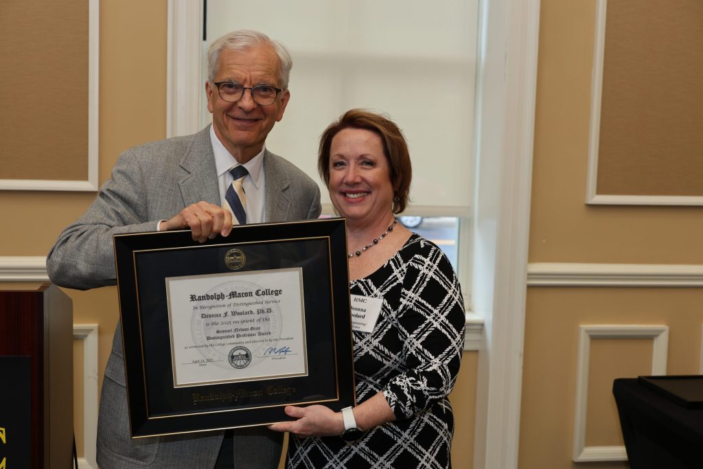An older man in a light suit and a woman in a patterned dress hold a framed certificate together and smile at the camera in a formal indoor setting.