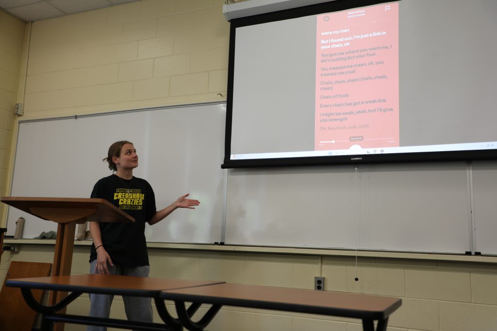 A presenter gestures toward a projector screen displaying song lyrics in red and white text in a classroom.