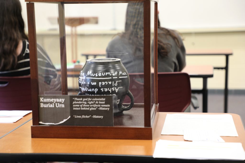 A Kumeyaay burial urn displayed in a glass case on a classroom table with students in the background.