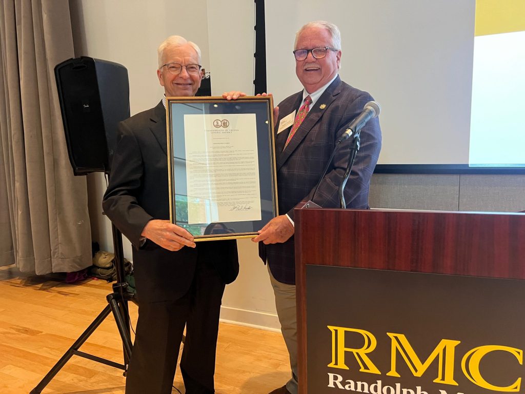 Two people stand beside a podium labeled "RMC," holding a framed document at a formal indoor event.