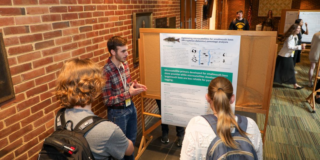 A person presents a scientific poster to three others in a hallway