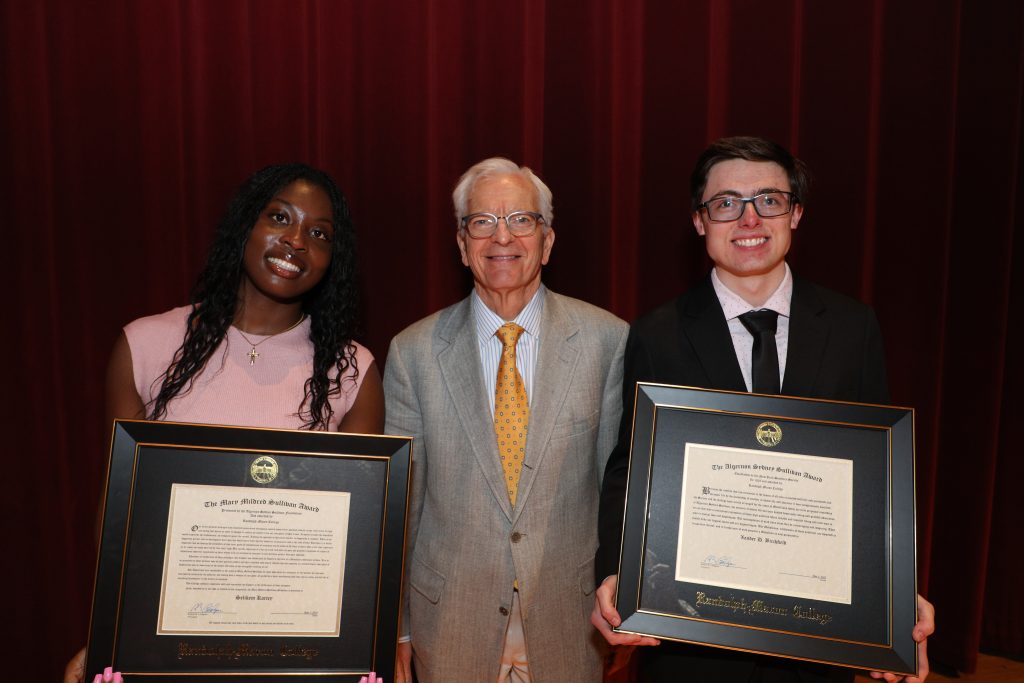 Three people posing for a photo; the two on either side hold framed certificates. They are in front of a red curtain.