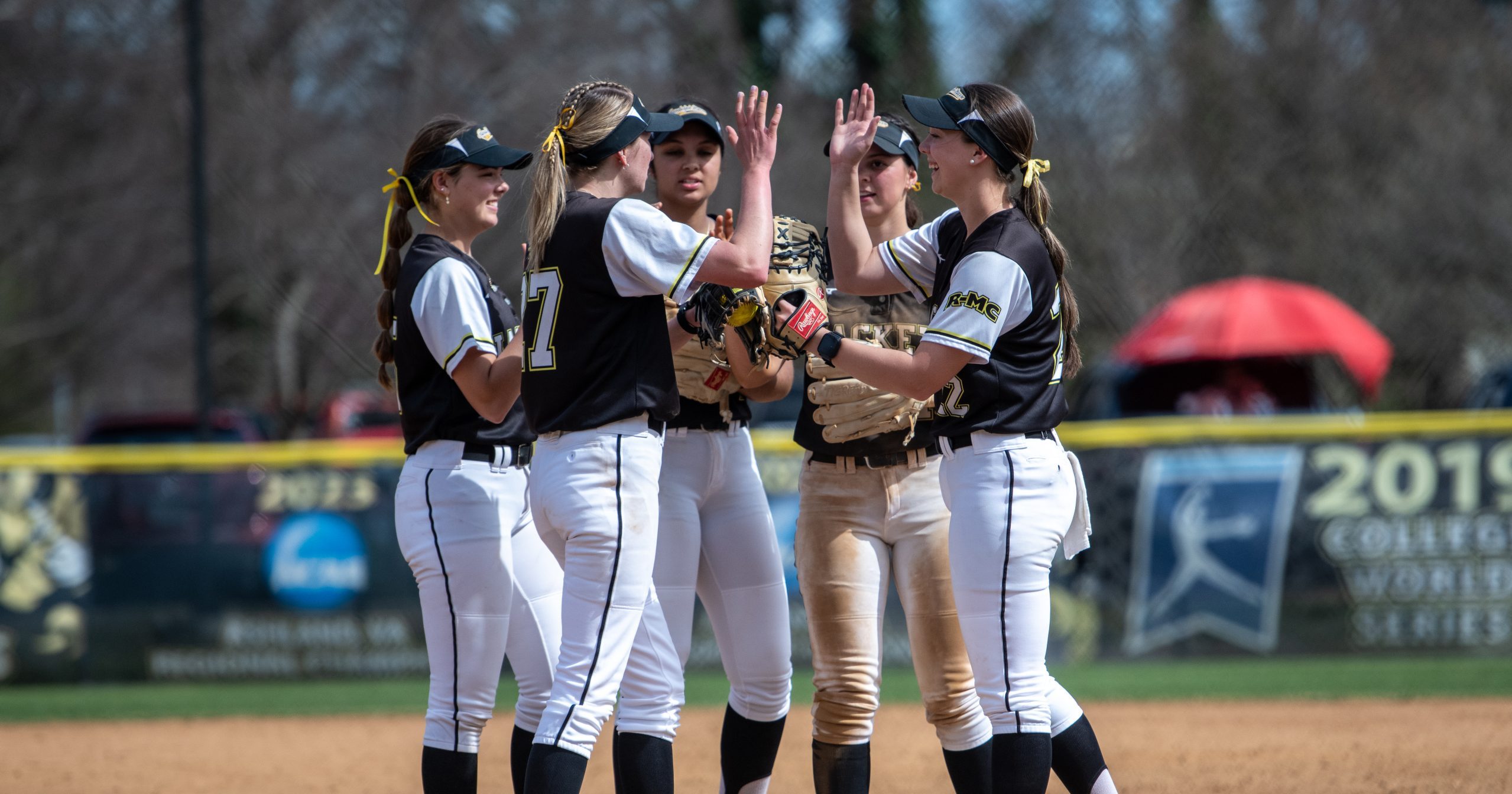Five softball players in matching uniforms stand on the field, high-fiving and talking during a game.