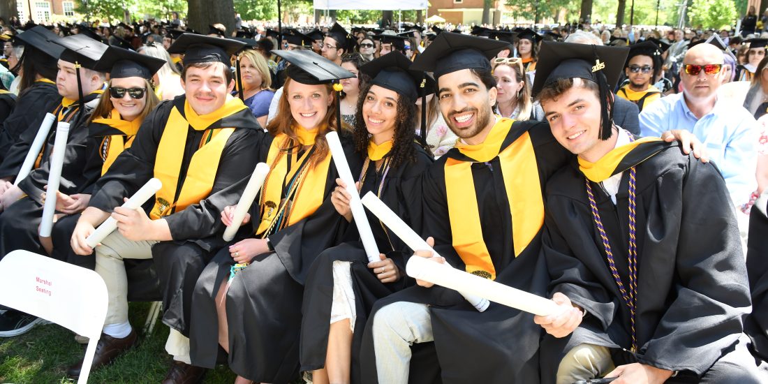 Graduates in caps and gowns sit together outdoors, holding diplomas, with a crowd in the background.