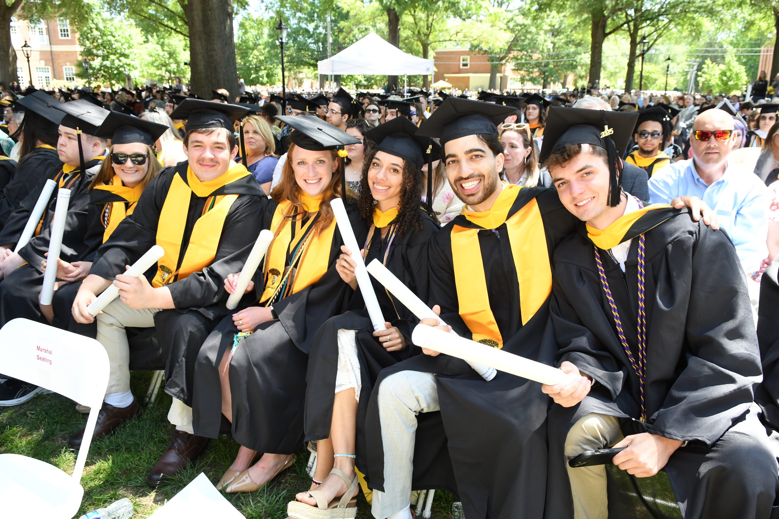 Graduates in caps and gowns sit together outdoors, holding diplomas, with a crowd in the background.