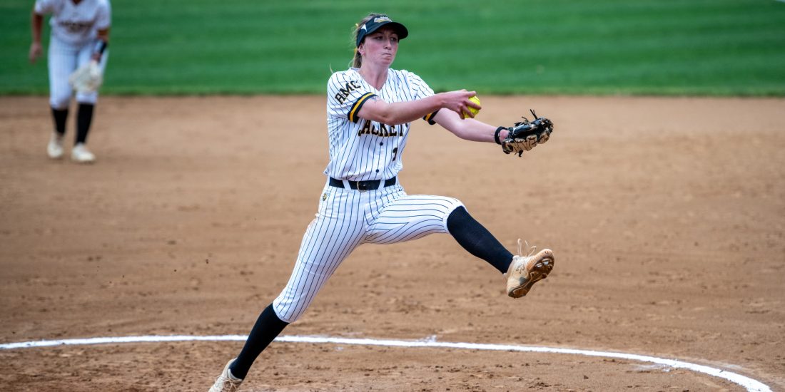 A softball pitcher delivers a pitch from the mound while an infielder stands ready.