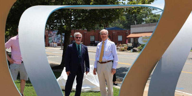 Two people in business attire stand outside near a large abstract metal sculpture, with a street and buildings in the background.