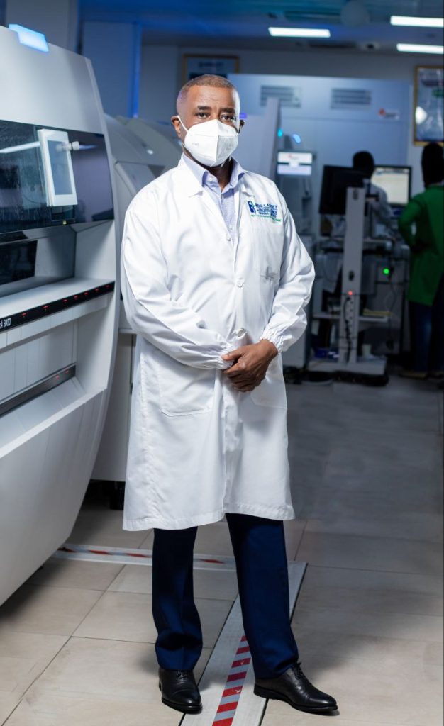 A person in a lab coat and face mask stands in a laboratory with medical equipment in the background.