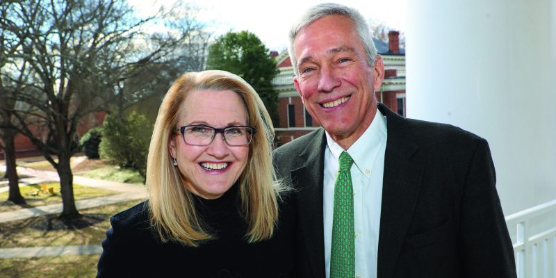 Two people stand side by side outdoors near a white column, with trees and a brick building in the background.