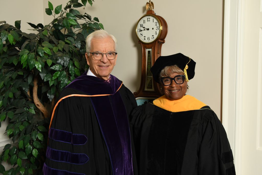 Two individuals in academic regalia stand indoors, smiling at the camera, with a wall clock and potted plant in the background.
