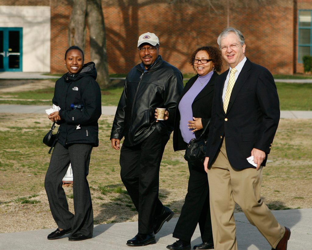 Four people walk together on a sidewalk near a building, with trees and grass in the background.