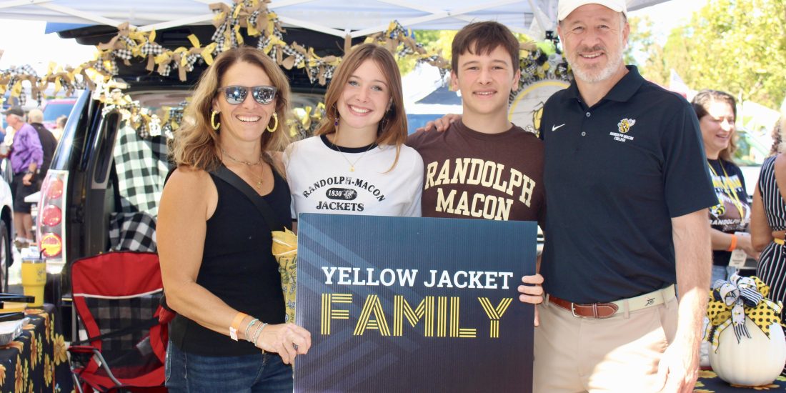 Four individuals stand under a decorated tent at a Randolph-Macon College Family Weekend event, holding a sign that reads "Yellow Jacket Family.