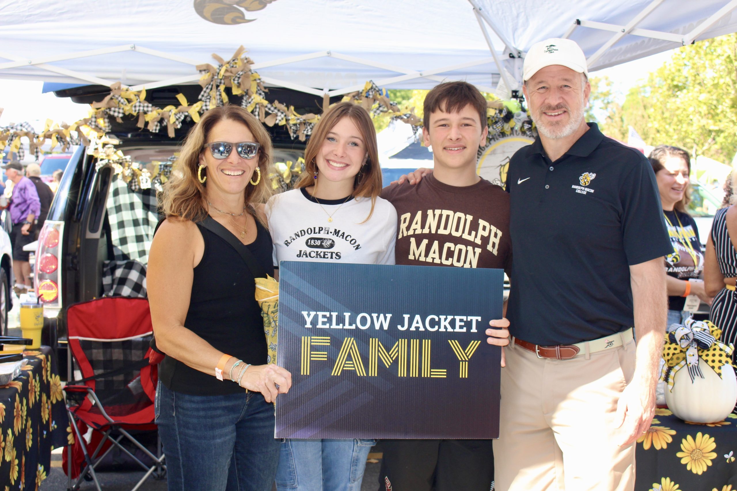 Four individuals stand under a decorated tent at a Randolph-Macon College Family Weekend event, holding a sign that reads "Yellow Jacket Family.