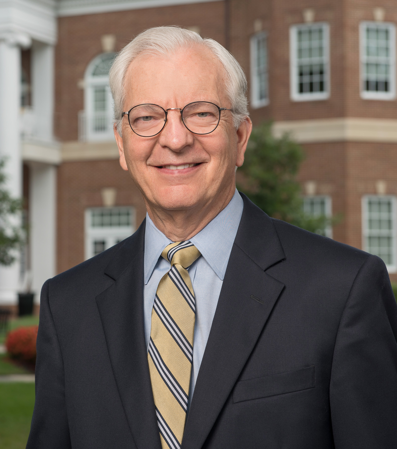 A person in formal attire stands outdoors in front of a red brick building with white trim and multiple windows.