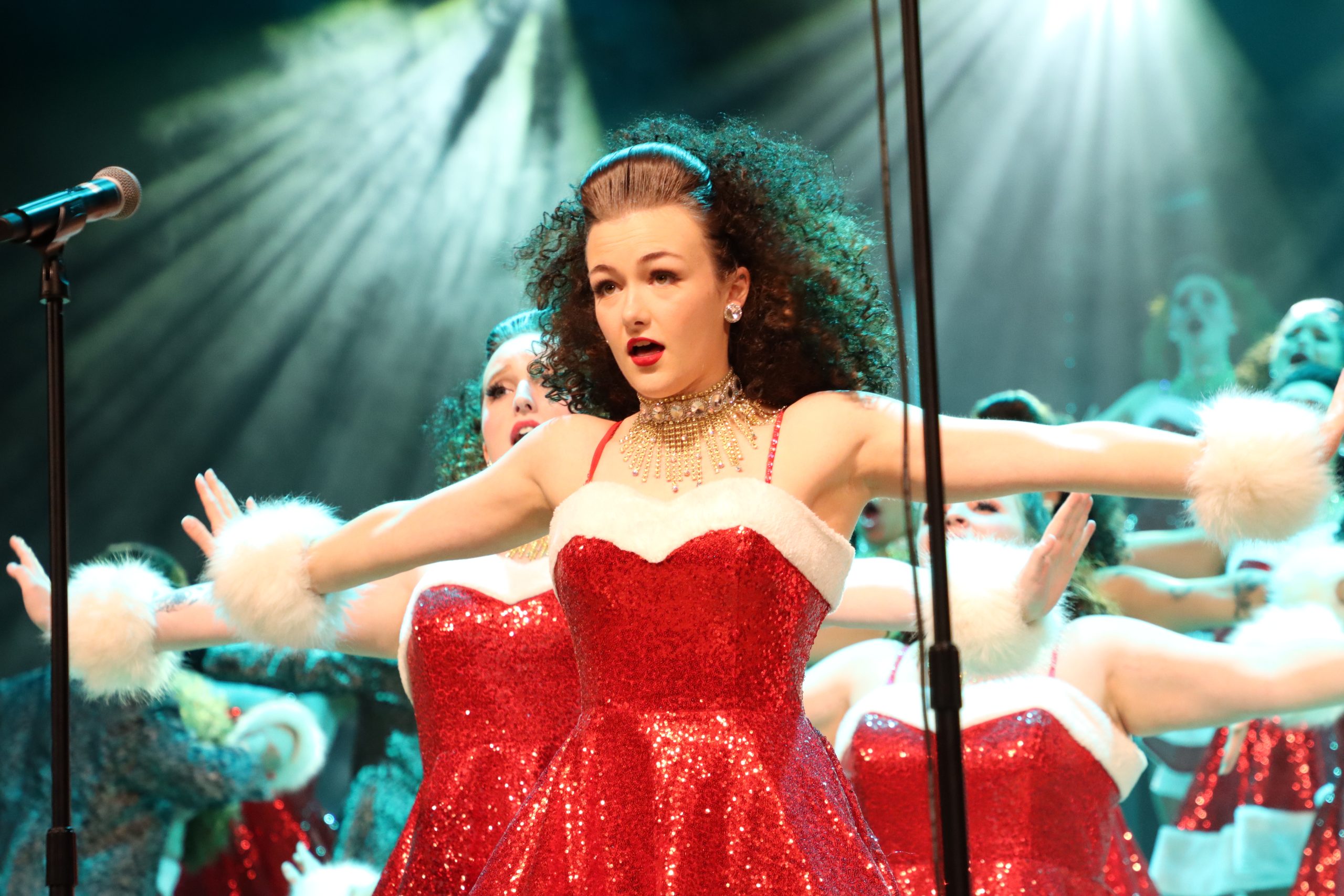 Performer in a sequined dress on stage with backup dancers, arms outstretched under bright stage lights.