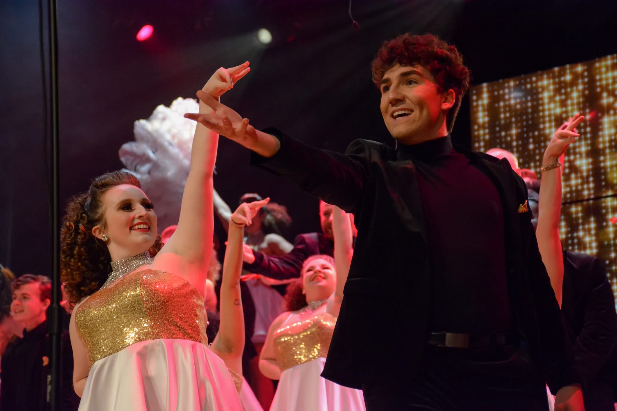 A group of performers in formal attire dance energetically on stage under colorful lighting.