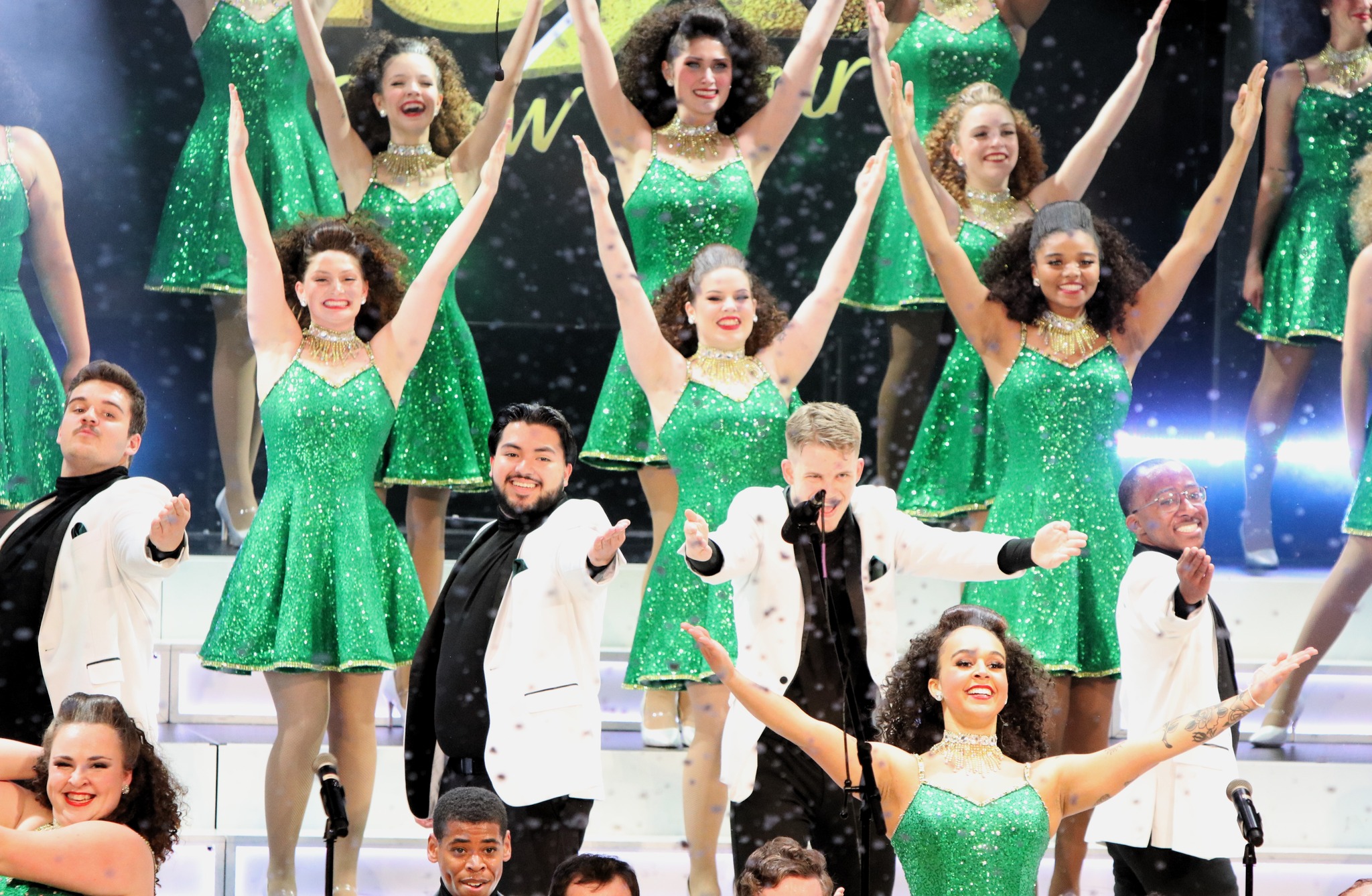 A group of performers in coordinated green and white costumes sing and dance on stage, striking dynamic poses.