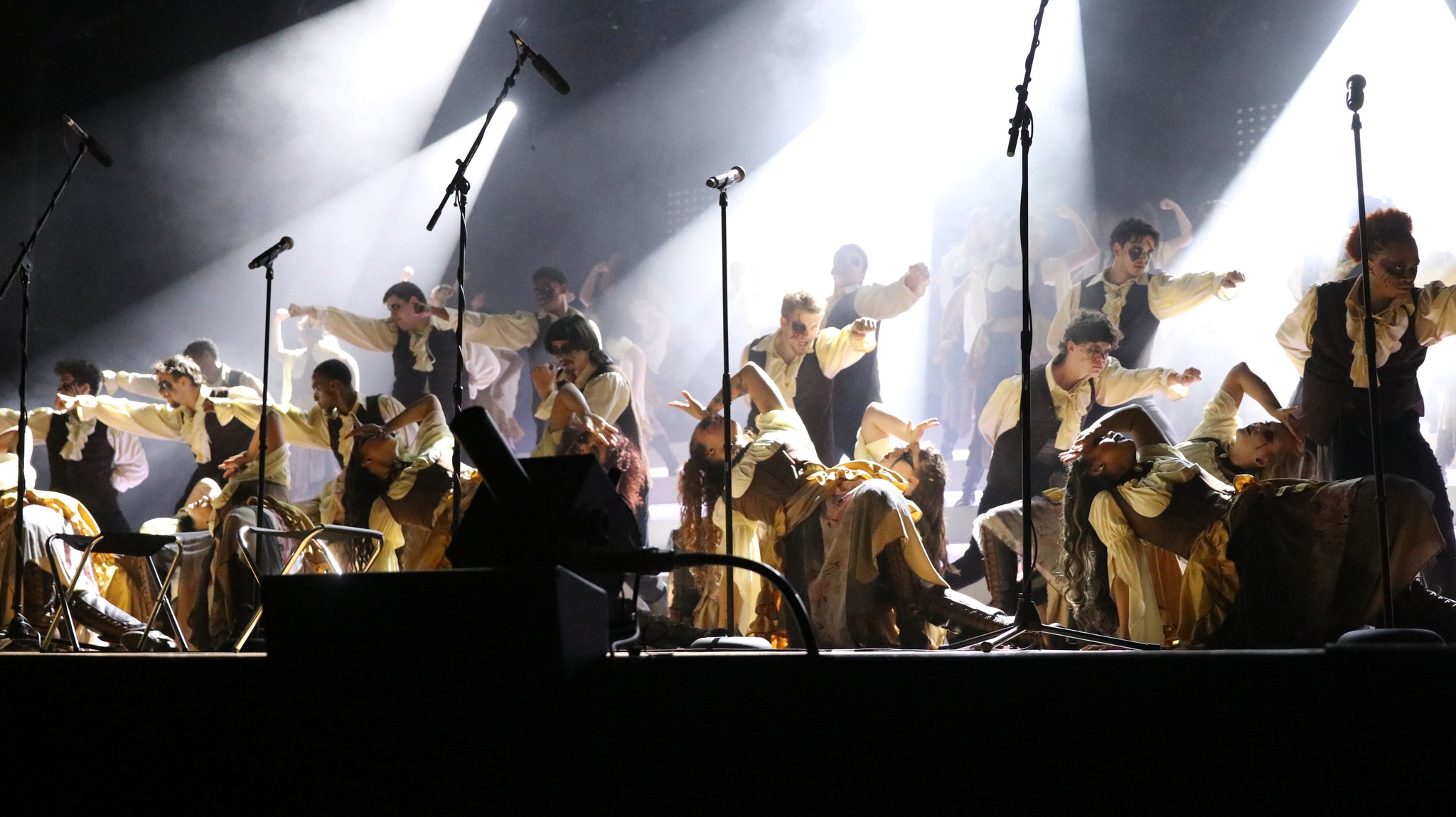 A group of performers in matching costumes pose on stage under bright spotlights with microphones in front of them.