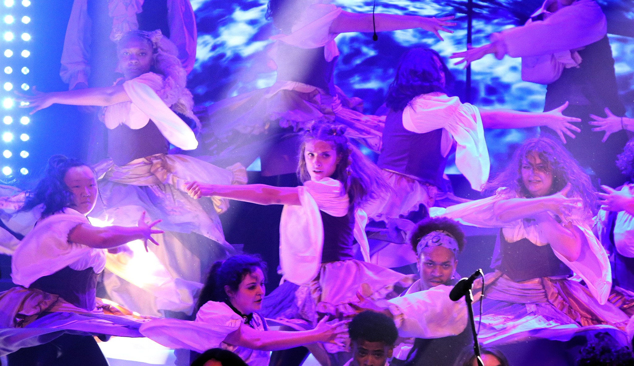A group of performers in costume dance on stage with dramatic lighting and blue visual effects in the background.
