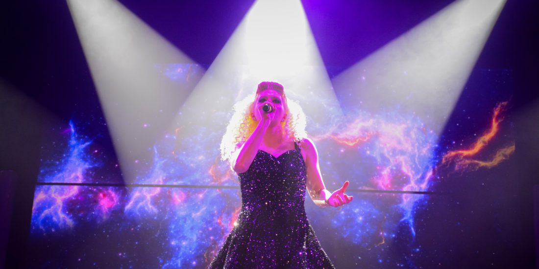 A performer sings on stage under bright lights, with a colorful cosmic background displayed behind them.